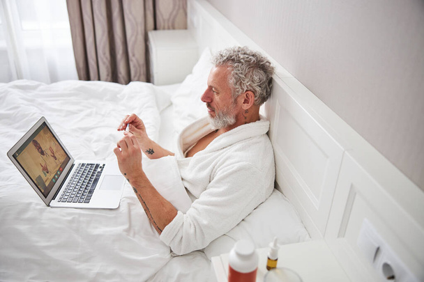 Adult Caucasian man in white bathrobe talking with physician online while sitting on modern interior room - Valokuva, kuva