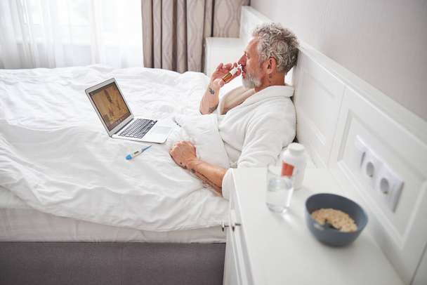 Varón caucásico barbudo sentado en la cama mientras habla con el médico en videollamada - Foto, Imagen