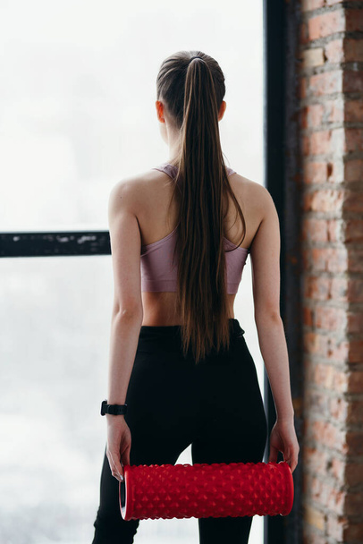 Photo from behind. A lean physique girl in sportswear holds a fascia in her hands while standing against a background of windows - Zdjęcie, obraz