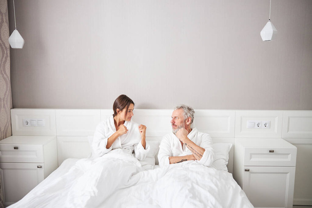 Angry married couple in white bathrobes looking to each other while gesturing in home apartment - 写真・画像