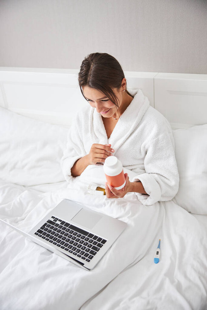 Charming elegant caucasian woman talking online on the internet while holding pill jar in hands - Foto, imagen