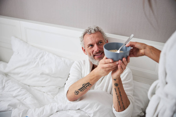Handsome adult man looking away while holding bowl with cereal in home modern interior room - 写真・画像