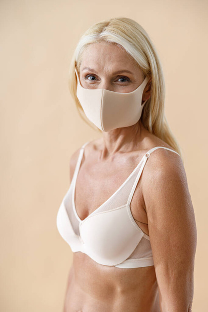 Portrait of beautiful caucasian mature blonde woman in underwear and protective face mask looking at camera while posing half naked against beige background - Φωτογραφία, εικόνα