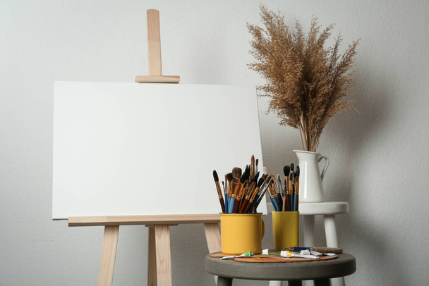 Easel with blank canvas, brushes, paints and palette in studio - Foto, imagen
