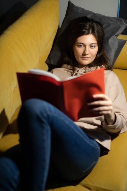 Vonzó hosszú hajú lány szándékosan olvas könyvet fekvő kényelmes kanapé térd hajlított - Fotó, kép