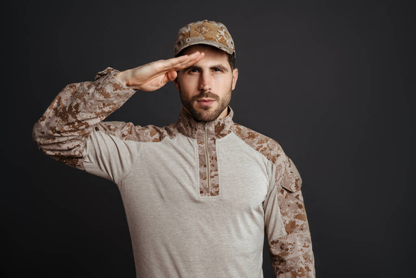 Confident masculine military man saluting and looking at camera isolated over black background - Photo, image