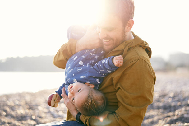Usmívající se otec v jasném slunečním světle objímá malé dítě v modré kombinéze s jablkem v ruce na hrudi. Detailní záběr - Fotografie, Obrázek