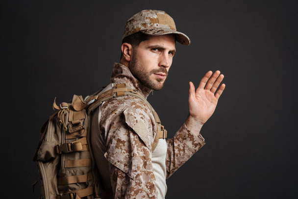 Zelfverzekerde mannelijke militaire man zwaaiende hand terwijl poseren met rugzak geïsoleerd over zwarte achtergrond - Foto, afbeelding