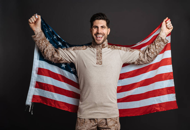 Happy masculine military man smiling while posing with american flag isolated over black background - Zdjęcie, obraz
