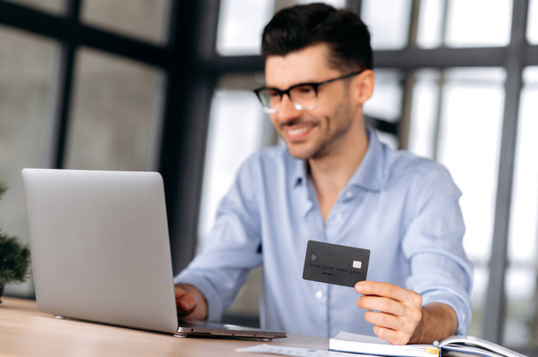 Blurred young modern Caucasian man wearing glasses, in casual clothes, sitting at table, using laptop and credit card to pay for online purchases or deliver goods home, smiling. Online shopping - Fotó, kép