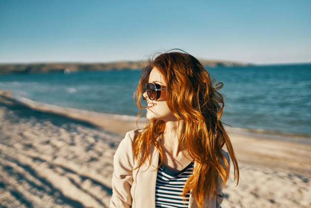 femme rousse sur la plage portant des lunettes près de la mer beige manteau t-shirt modèle - Photo, image