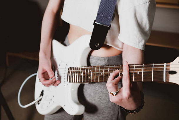 Γυναίκα καλλιτέχνης παίζει ηλεκτρική κιθάρα παίζοντας ένα τραγούδι. θέα καλλιεργειών. Μια γυναίκα με άσπρη κιθάρα που κάνει πρόβες στο σπίτι. μουσική ως χόμπι και δημιουργική αναψυχή, επιλεκτική εστίαση. - Φωτογραφία, εικόνα