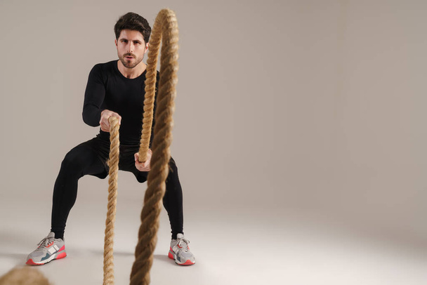 Handsome athletic sportsman working out with battle ropes isolated over grey background - 写真・画像