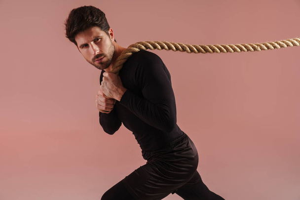 Confident young brunette sportsman pulling rope over pink wall background - Foto, immagini