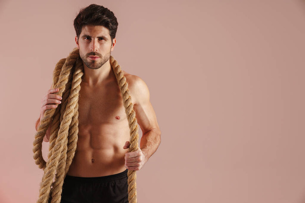 Serious young brunette shirtless man standing with rope over his shoulders over pink wall - Photo, image