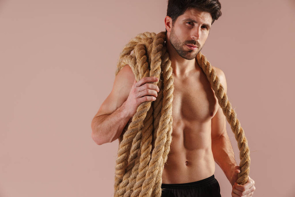 Serious young brunette shirtless man standing with rope over his shoulders over pink wall - Foto, afbeelding