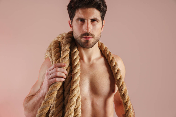 Serious young brunette shirtless man standing with rope over his shoulders over pink wall - Foto, Imagen