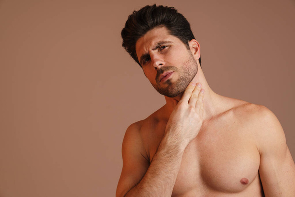 Brooding half-naked man looking at camera and rubbing his neck isolated over beige background - Foto, afbeelding