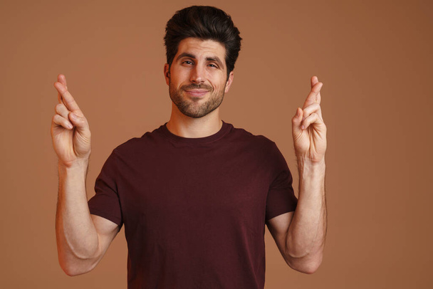 Confused masculine man posing with fingers crossed for good luck isolated over beige background - Foto, immagini