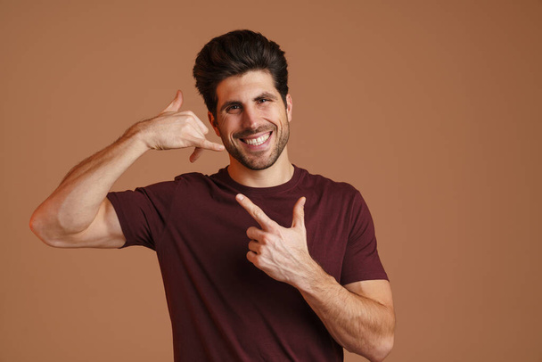 Happy man pointing finger upward while showing handset gesture isolated over beige background - Foto, imagen
