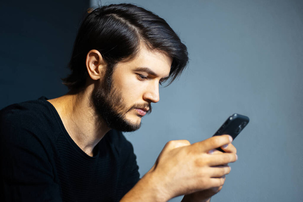 Portrait of young man using smartphone on the background of grey and black wall. - Photo, Image