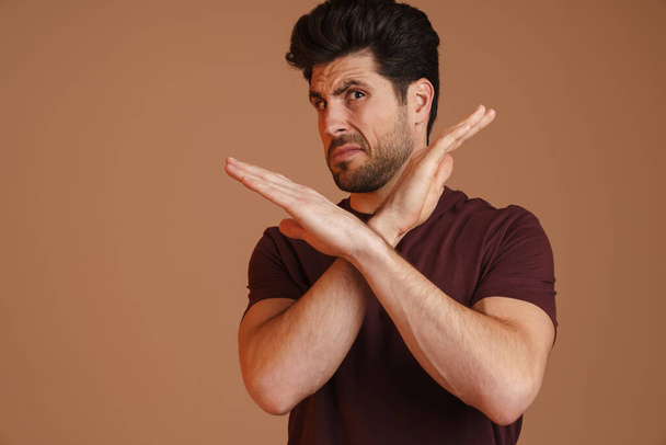 Displeased unshaven young man showing stop gesture at camera isolated over beige background - Фото, изображение