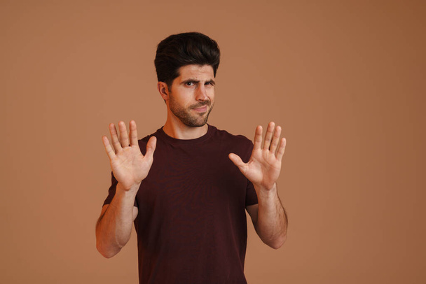 Displeased unshaven young man showing stop gesture at camera isolated over beige background - Zdjęcie, obraz