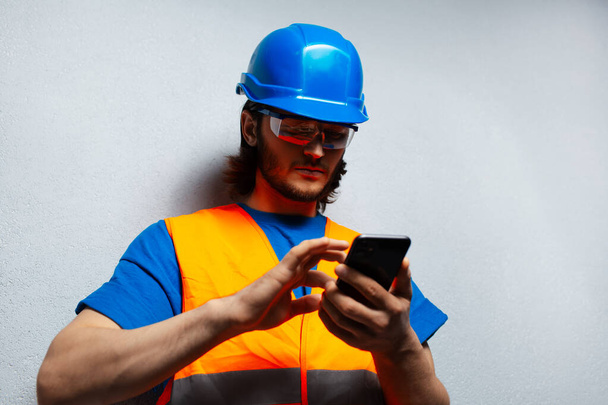 Close-up portrait of young man, construction worker engineer, using smartphone, wearing safety equipment on the background of grey textured wall. - Photo, Image