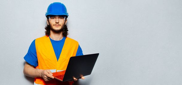 Portrait of young confident man, construction worker engineer wearing safety equipment, using laptop on grey background. Panoramic banner view with copy space. - Foto, imagen