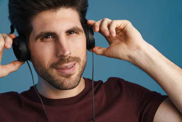 Pleased handsome man listening music with headphones isolated over blue background - Foto, imagen