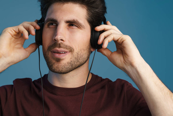 Pleased handsome man listening music with headphones isolated over blue background - Foto, afbeelding