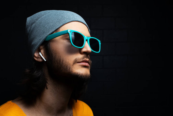 Studio portrait of young serious man with wireless earphone in ear, wearing sunglasses and hat on black background. - Foto, imagen
