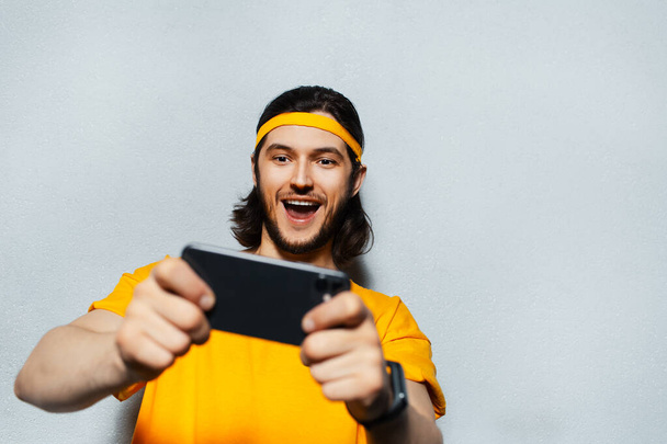 Studio portret młodego człowieka szczęścia przy użyciu smartfona na szarym tle teksturowanym; noszenie żółtej opaski na głowę i koszulę. - Zdjęcie, obraz