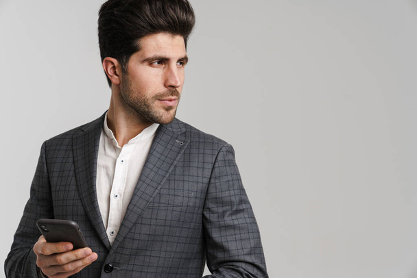 Confident young man in jacket posing and using mobile phone isolated over grey background - Φωτογραφία, εικόνα