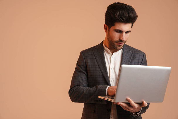 Serious young man in jacket standing while working with laptop isolated over beige background - Фото, изображение