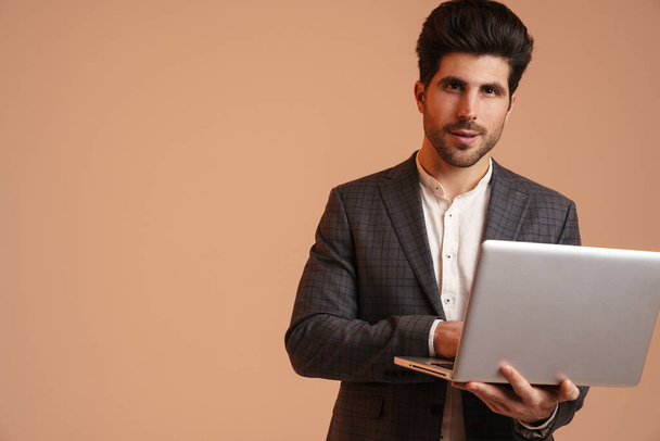 Serious young man in jacket standing while working with laptop isolated over beige background - Foto, Imagem