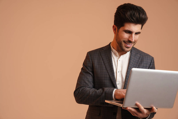 Smiling young man in jacket standing while working with laptop isolated over beige background - Φωτογραφία, εικόνα
