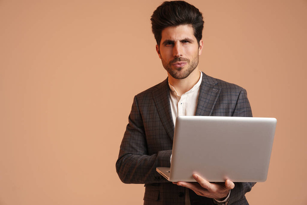 Serious young man in jacket standing while working with laptop isolated over beige background - Φωτογραφία, εικόνα