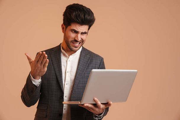 Elégedetlen jóképű férfi gesztikulál, miközben dolgozik laptop elszigetelt bézs háttér - Fotó, kép