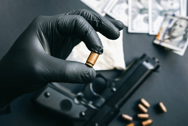 Arma tendida sobre la mesa. Hombre con guantes negros sosteniendo balas. Venta ilegal de drogas. Problemas criminales. Dólares. - Foto, Imagen