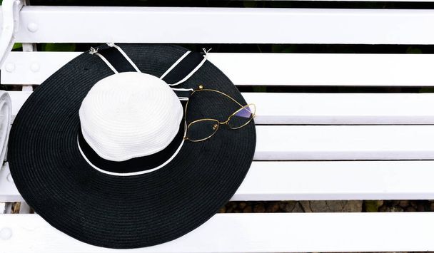 Cappello moda donna bianco e nero con occhiali da vista dorati su una panchina bianca. - Foto, immagini