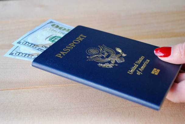 Ženské prsty držící americký pas s dolarovými bankovkami na dřevěném pozadí. Cestovní koncept. - Fotografie, Obrázek