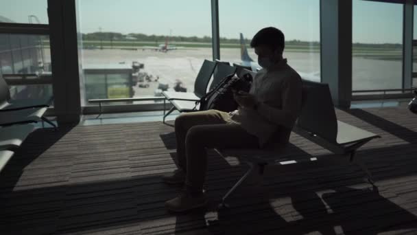 Ο μασκοφόρος κάθεται στο αεροδρόμιο, σε θέση κοινωνικής απόστασης, δίπλα στο παράθυρο, πριν επιβιβαστεί στο αεροπλάνο και χρησιμοποιεί το τηλέφωνό του από πλήξη. Καθυστέρηση πτήσης. Ακύρωση πτήσεων λόγω καραντίνας και αποκλεισμού covid 19 - Πλάνα, βίντεο