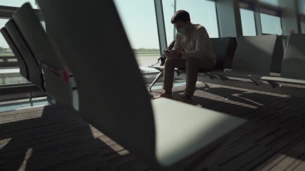 Νέα κανονική έννοια για τα αεροπορικά ταξίδια. Κοινωνική απόσταση στο αεροδρόμιο. Ένας άνδρας που φοράει μάσκα covid 19 χρησιμοποιώντας το smartphone του κάθεται στο λόμπι πριν από την επιβίβαση. Ταξιδεύοντας μόνος κατά τη διάρκεια του coronavirus - Πλάνα, βίντεο