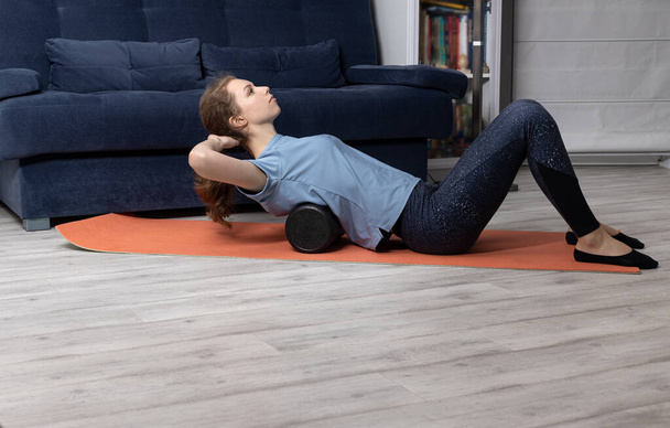 oefeningen voor de rug met een gymnastiekroller op de mat. Meisje is bezig met stretching thuis op de vloer. - Foto, afbeelding