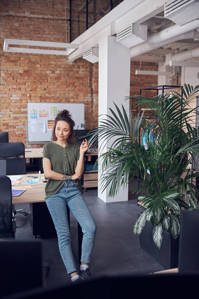 Full length shot of interior designer woman holding glasses and looking away while standing in modern green office on a daytime and thinking about new project - Photo, Image