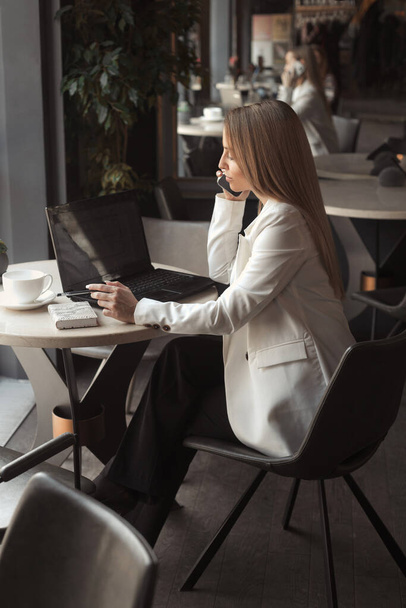 Business woman drink coffee in a white jacket and working with laptop in cafe with dark black interior - Foto, immagini