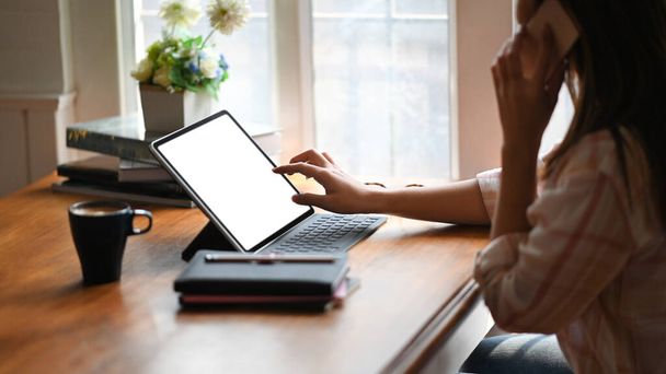 Boczny obraz pięknej kobiety w pasiastej koszuli dzwoniącej przez smartfona dotykając białego tabletu z klawiaturą i siedząc przy drewnianym stole z oknami jako tłem. - Zdjęcie, obraz