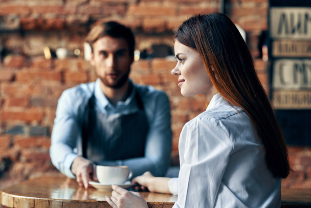 mujer feliz con una taza de café y camarero masculino en el delantal en la mesa en el interior - Foto, Imagen