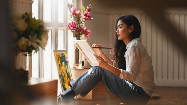 Resim sanatçısı genç bir kızın elinde fırça tutarken ve tuvale yağlı boya renkleri çizerken modern sanat stüdyosunda otururken. Sanatçı olarak yaratıcı kadın kavramı. - Fotoğraf, Görsel